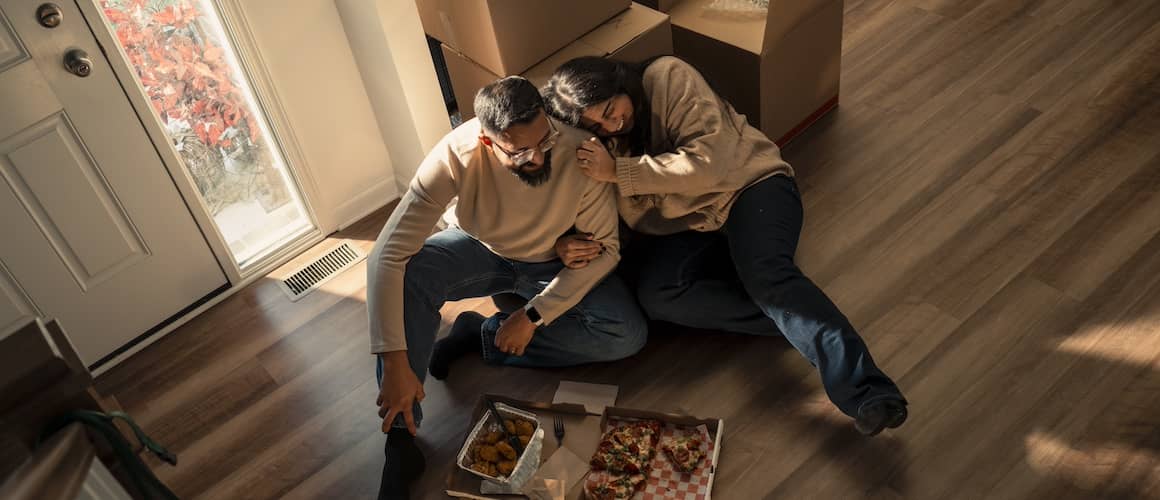 Couple snuggling and eating pizza on floor of new home surrounded by unopened moving boxes.