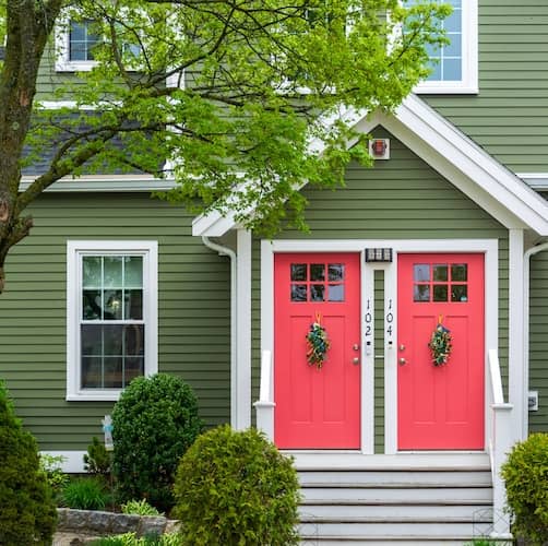 Duplex with green siding and pink doors. A large tree frames the front yard and shrubs flank the entrance staris.