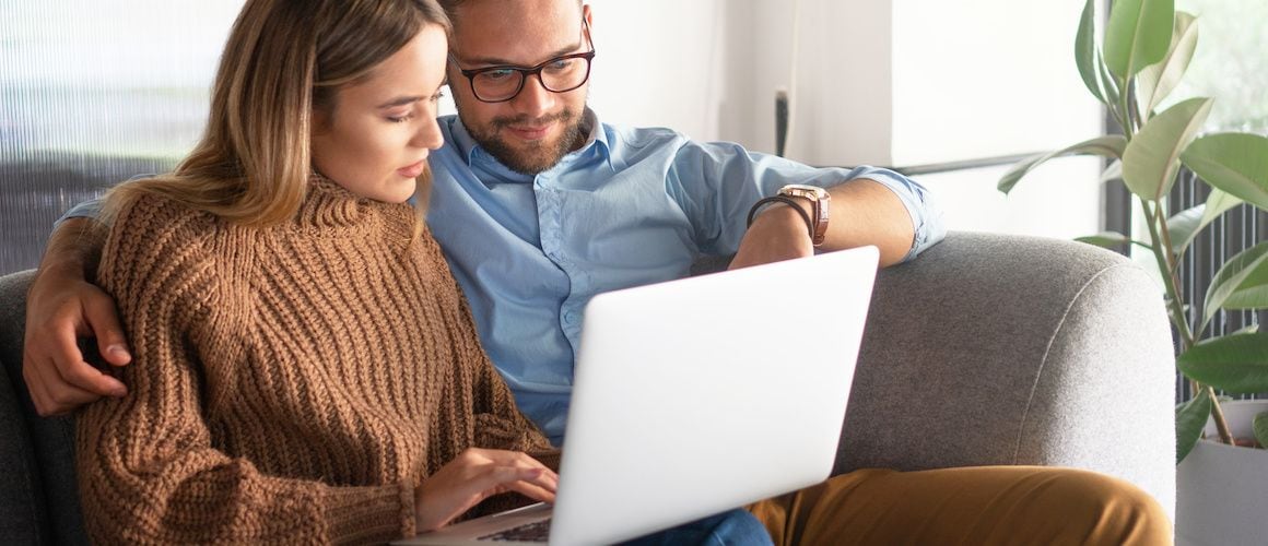 Smiling young couple using silver laptop while green houseplants sit on the wooden table behind them.