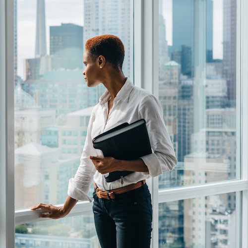 Realtor holding portfolio and looking out of floor to ceiling windows in commercial high rise with view of city skyline.