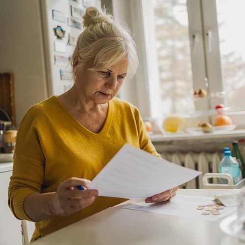 Senior woman wearing yellow filling out financial papers in her kitchen.