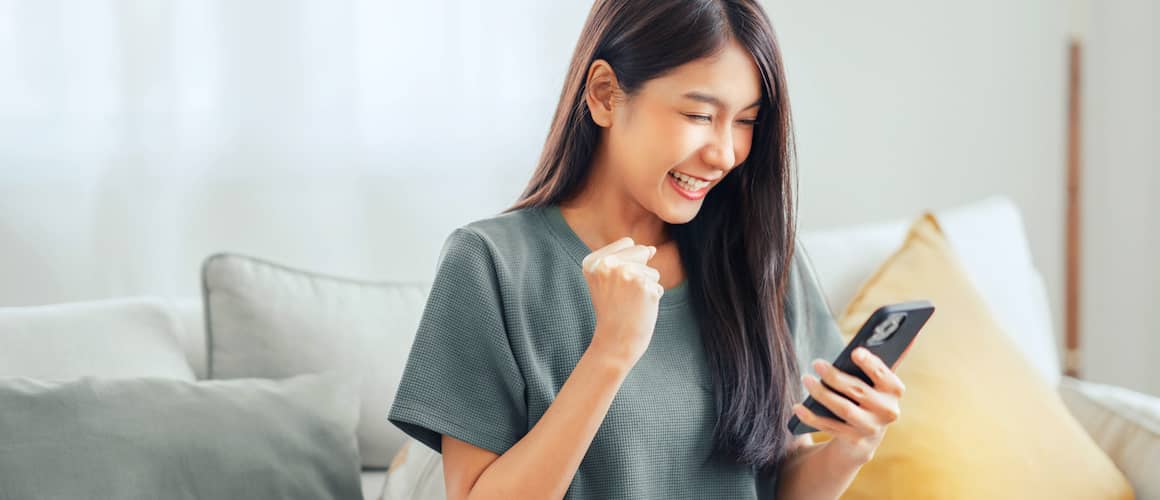 Young woman on her couch smiling and looking happily at her phone, with a fist pumping next to her in triumph.