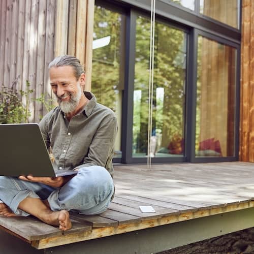 Man sitting cross legged with his laptop on his back deck of wood panelled home.