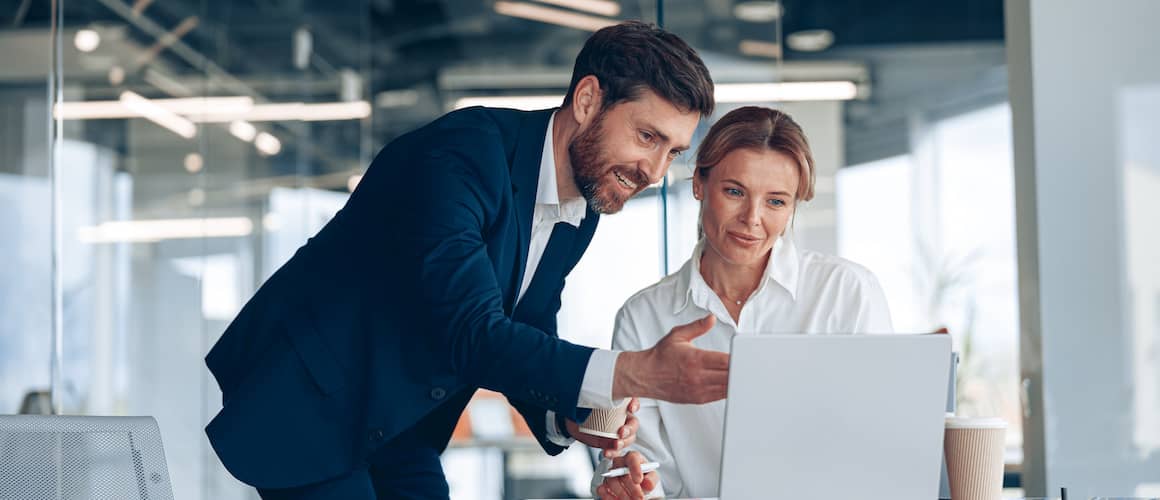 Man in business suit leaning over to point out something on the laptop screen of woman sitting at desk.