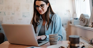 A woman with glasses wearing a denim shirt sits at her kitchen table looking at her laptop.