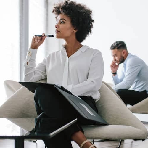 A woman sitting in a fancy room thinking something with a pen in hand while a man in background is talking on phone.