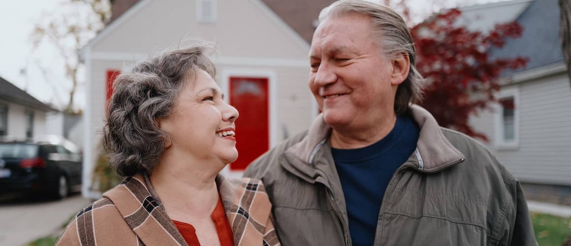 Older couple smiling at one another in front of home.