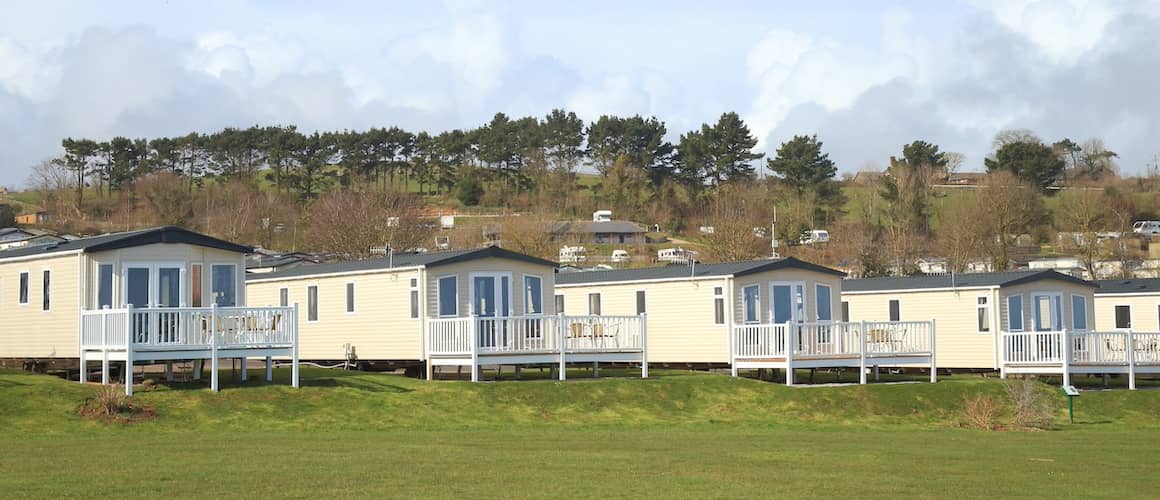 Row of white manufactured homes with expansive green lawn in foreground and trees in the background.