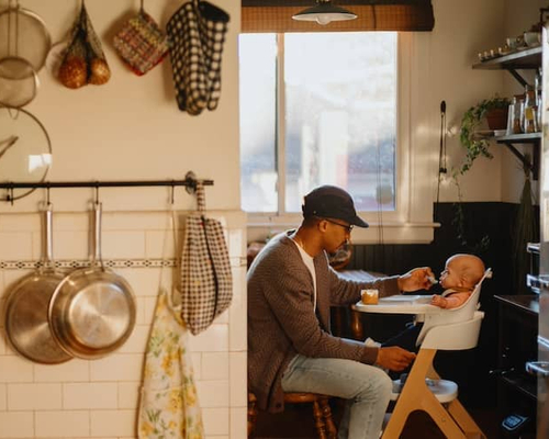 Mother in kitchen cooking while father feeds child in high chair in dining room.