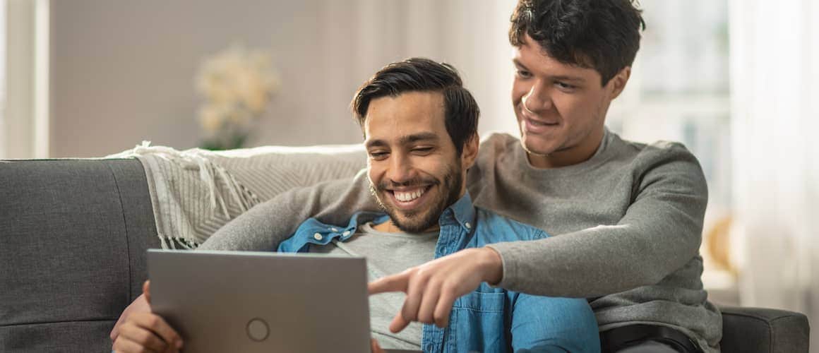 A gay couple cuddling on a couch while using a computer.