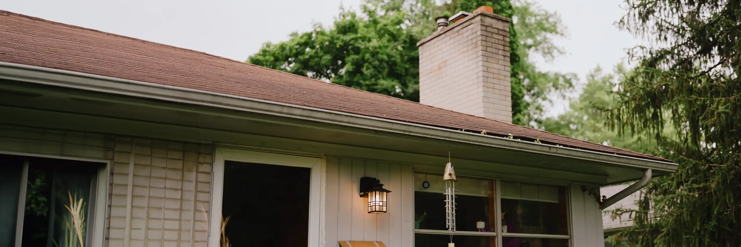 Partial exterior view of a welcoming single-story home with a red-brown roof, white brick chimney, and outdoor light fixture by the door, framed by green trees.