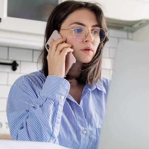 A woman sitting at the kitchen table looking at her computer and on the phone.