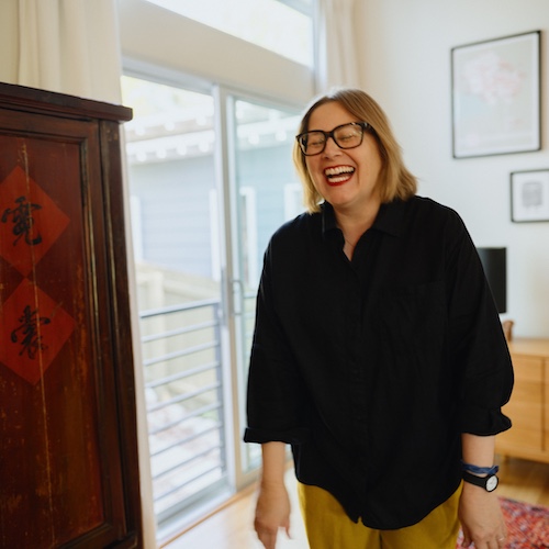 Woman in glasses next to dresser laughing.