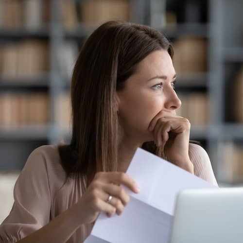 A woman lost in thought while holding a piece of paper in front of a computer screen.