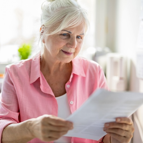 Senior woman in pink blouse reviewing paperwork in sunny living room.