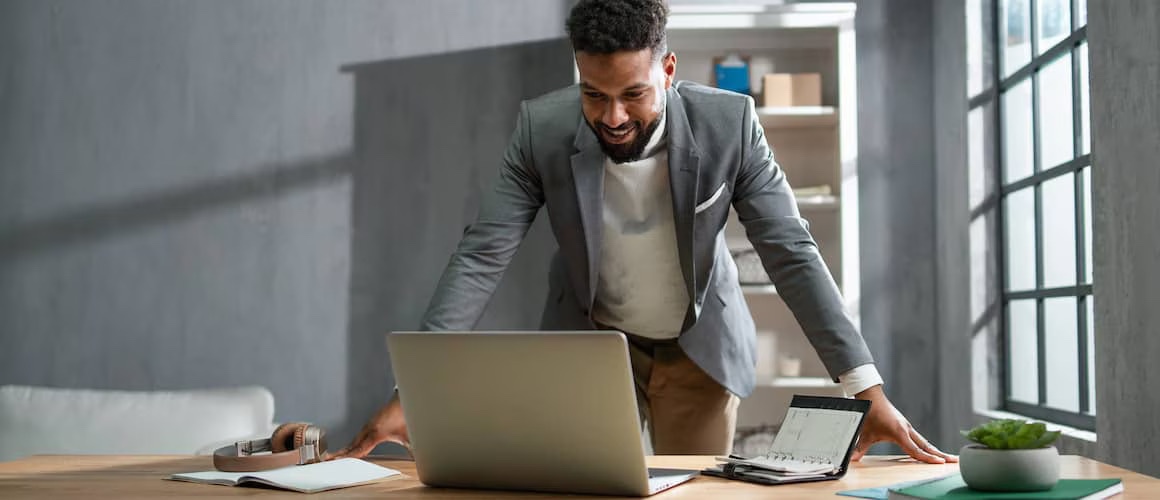 Young professional man standing at home office desk with open laptop.