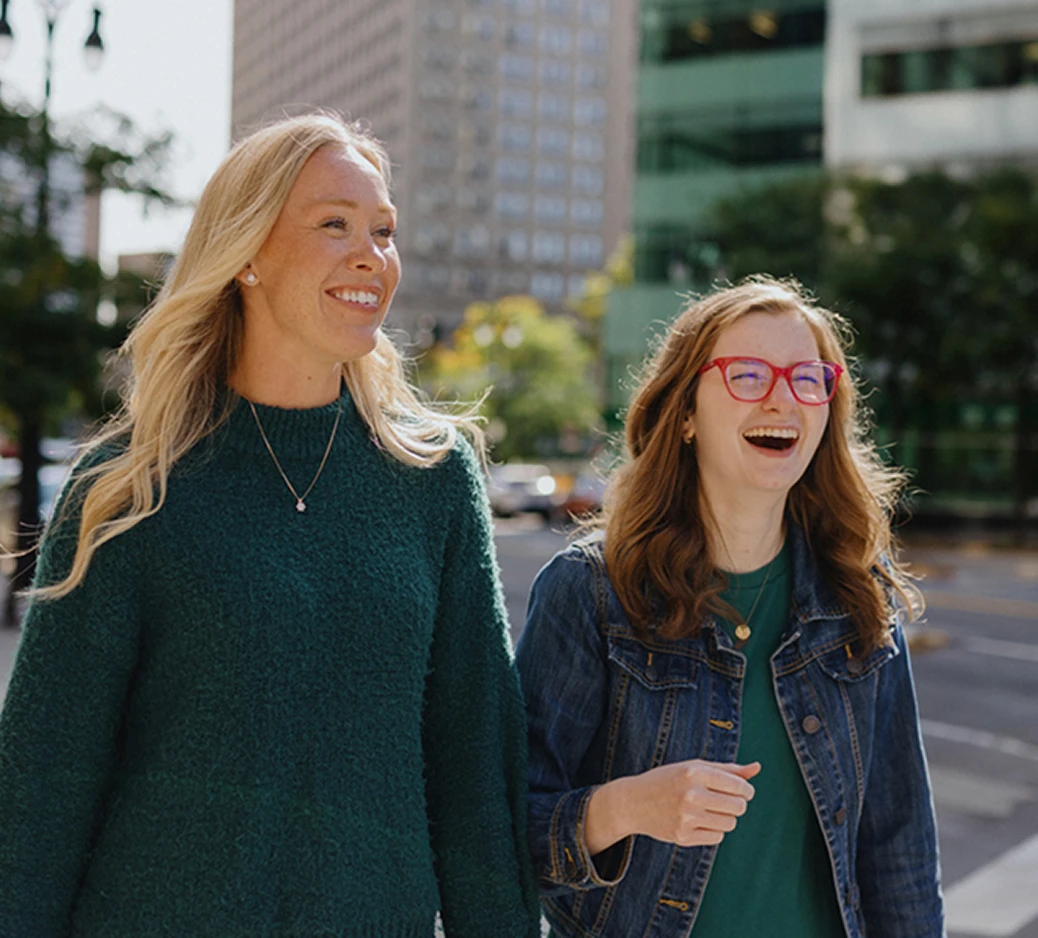 A person with short dark hair is seen from behind, wearing a white t-shirt with the text "HELP EVERYONE HOME" written on it in colorful letters. The person is holding a rainbow-colored umbrella and participating in a parade or march. Other people around are also holding rainbow flags and umbrellas, indicating support for LGBTQ+ pride. The event appears to be taking place on a city street with buildings and traffic lights visible in the background.