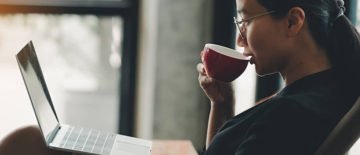 Woman staring at laptop while sipping a warm beverage in a red cup.