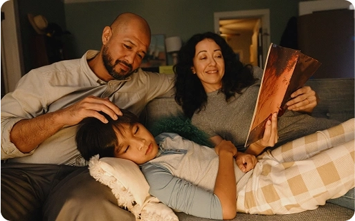 Two parents sitting on a couch with a child resting on a pillow, while the mother reads a book.