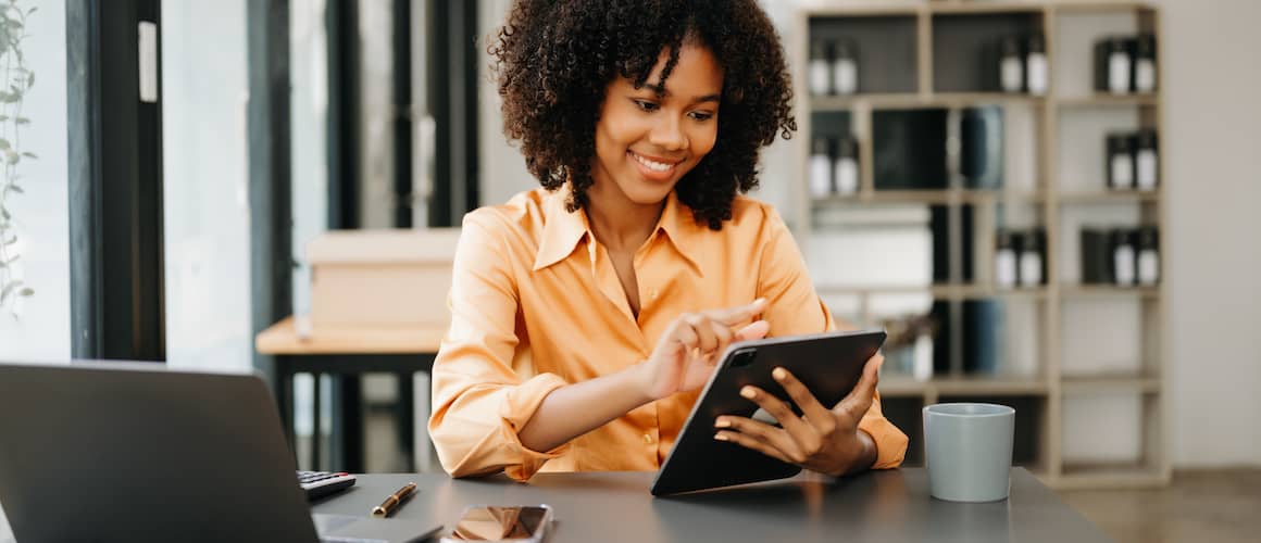 Woman smiling and working on a tablet, with a nearby cup of coffee and open laptop on her desk.