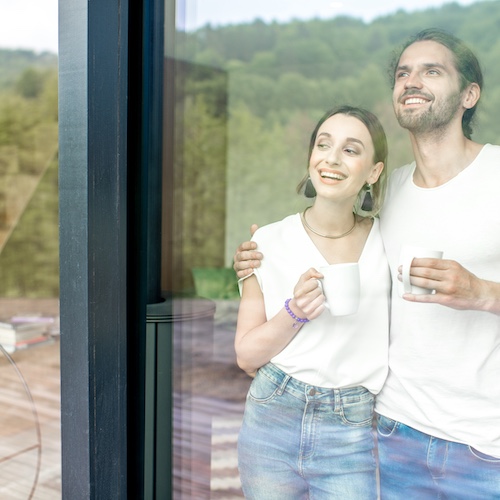 Young couple looking out sliding glass doors to expansive wooded hills in their home, drinking coffee.