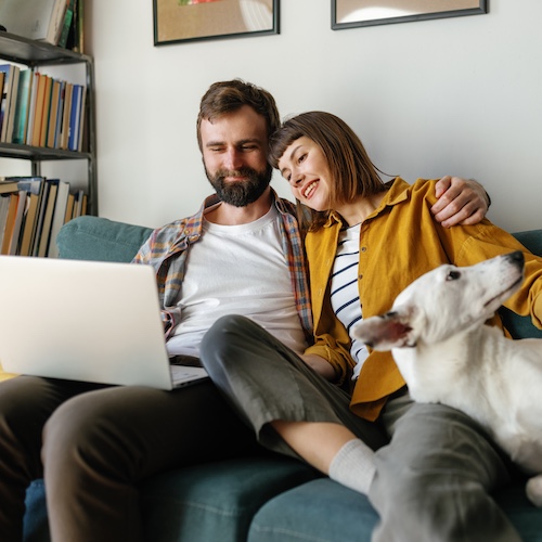 Young couple snuggling on couch with their dog, looking at information on a laptop.