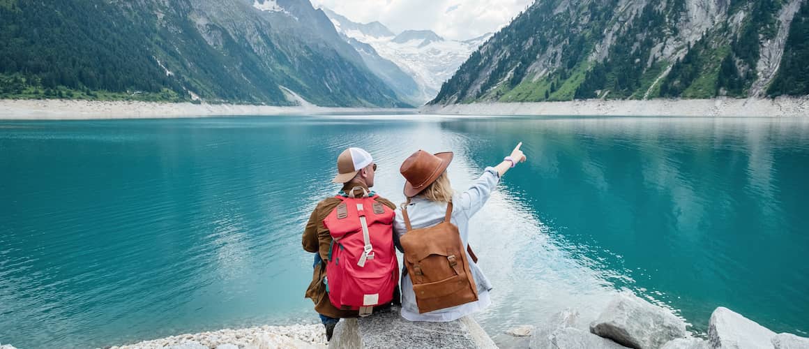 Man and woman sitting in front of lake and mountains pointing and looking in the distance.