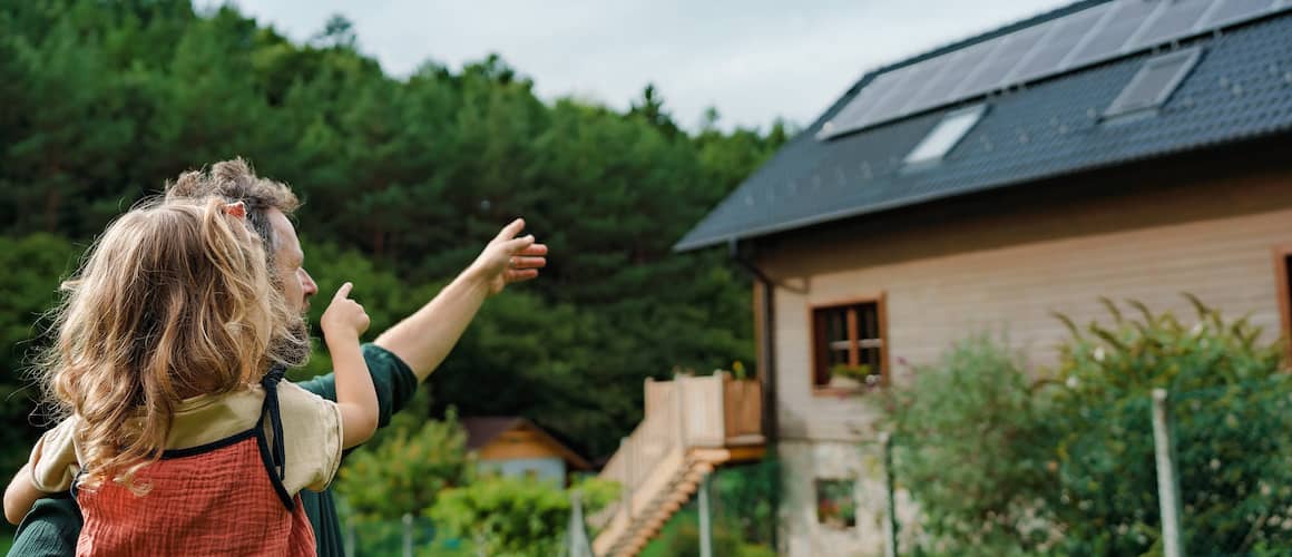 Father holding young daughter both pointing at a home with solar panels on its roof. It's a rustic style home with a lot of green trees around the home.