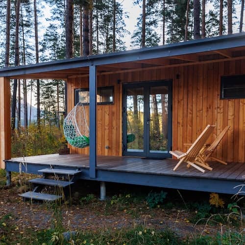 Wood panelled tiny home with covered porch with hanging chair surrounded by tall pine trees.