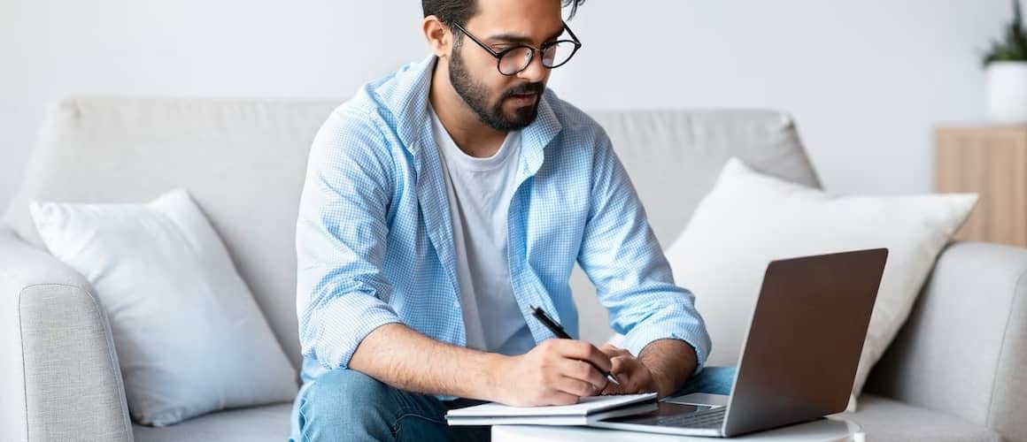 Man on couch viewing documents on laptop and writing on notebook.