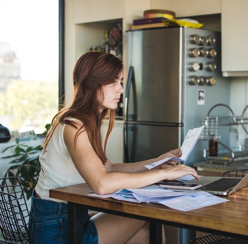 Young woman reviewing paperwork and documents online at kitchen table.