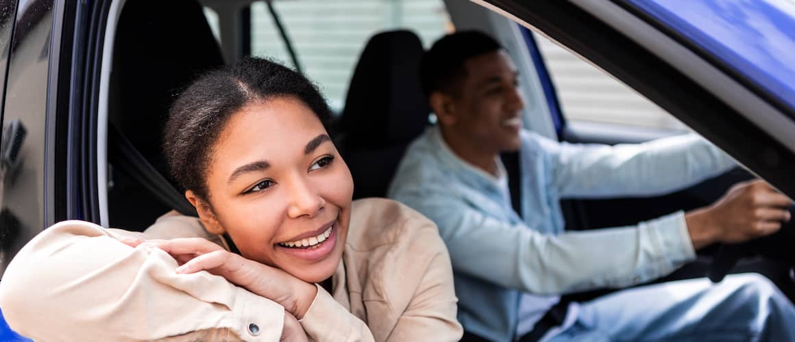 Couple in car together. Man is driving and smiling and young woman is leaning out of the window in passenger seat, smiling.