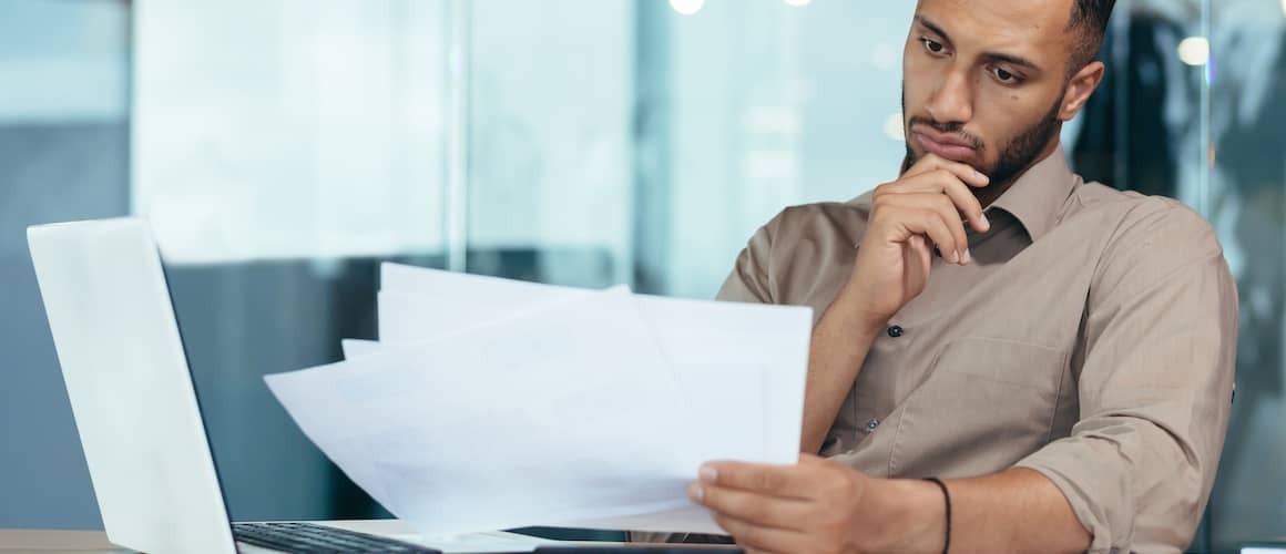 Man pensively looking at papers with open laptop nearby.