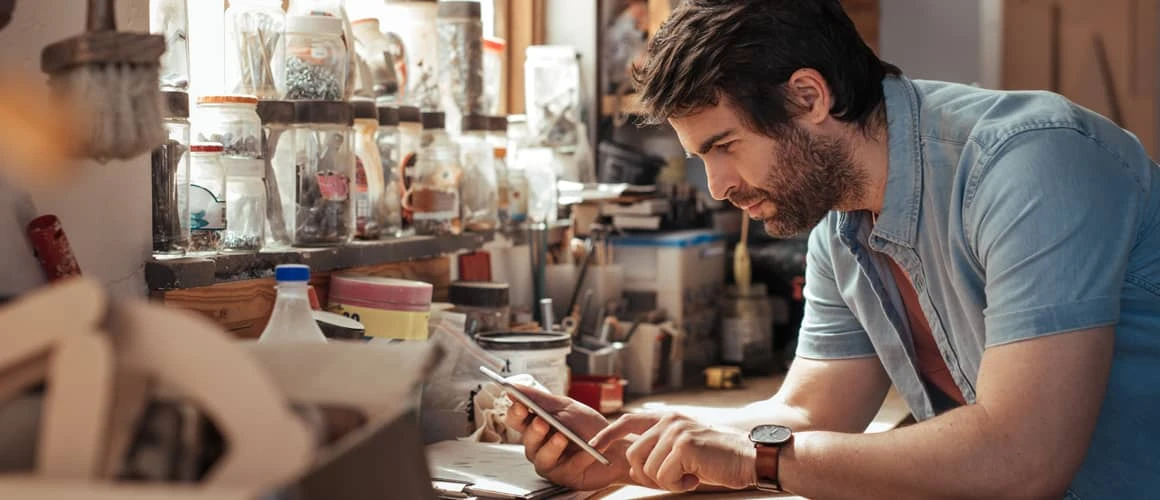 A white bearded man is looking intently at his smart phone while leaning on a workshop desk. He's holding the smart phone with his right hand, and is using his left hand to point at something on the screen or is scrolling down a web page.