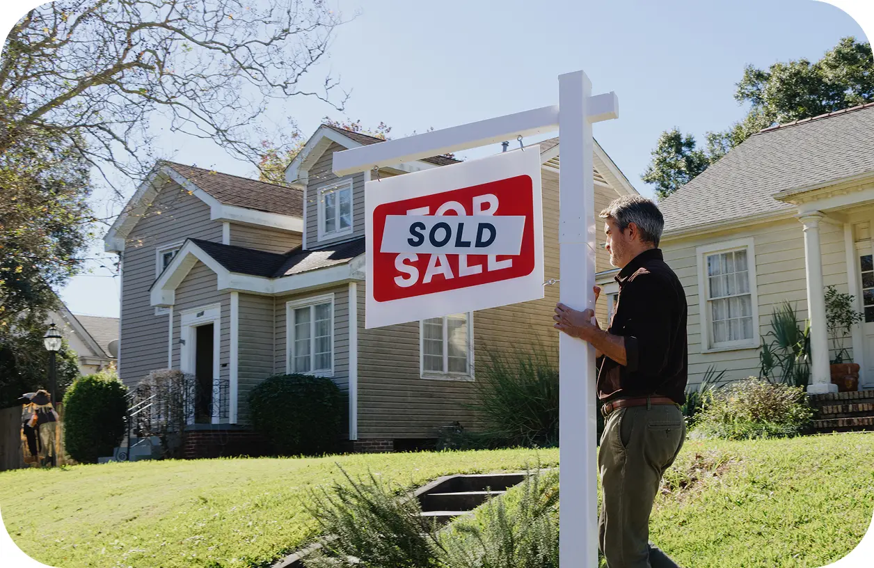 A man stands in the backyard of a modest home, carefully pulling a 'Sold' sign out of the grass.
