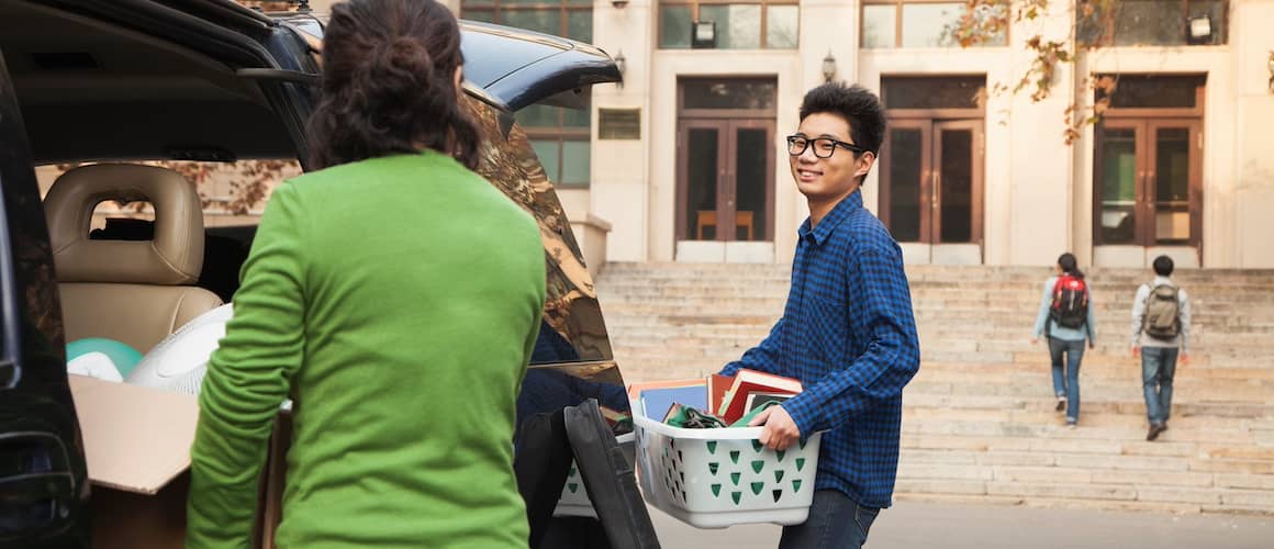 Mother helping son unpack car and carry laundry basket and boxes to dormitory.