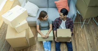 A couple sits on the floor of their new home surrounded by boxes.