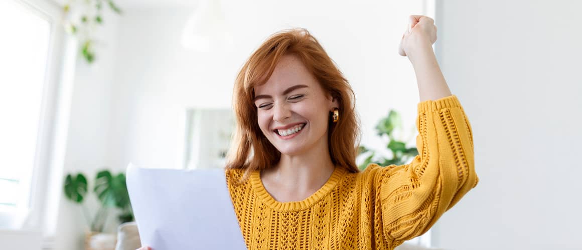 Woman sitting on couch holding a document with one hand and putting other hand in the air triumphantly.