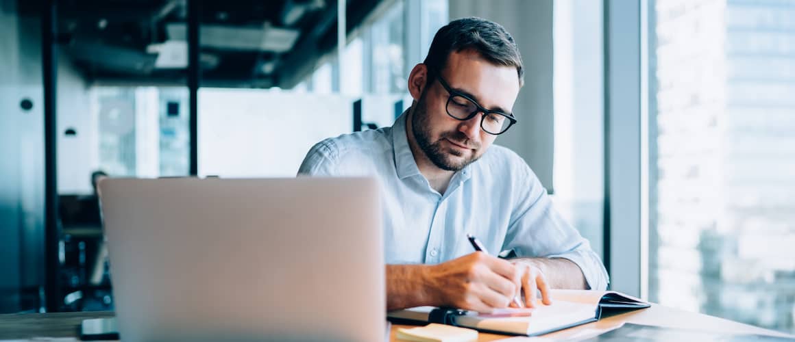 Young businessman sitting at his desk in his office while writing in a journal with his laptop open in front of him.