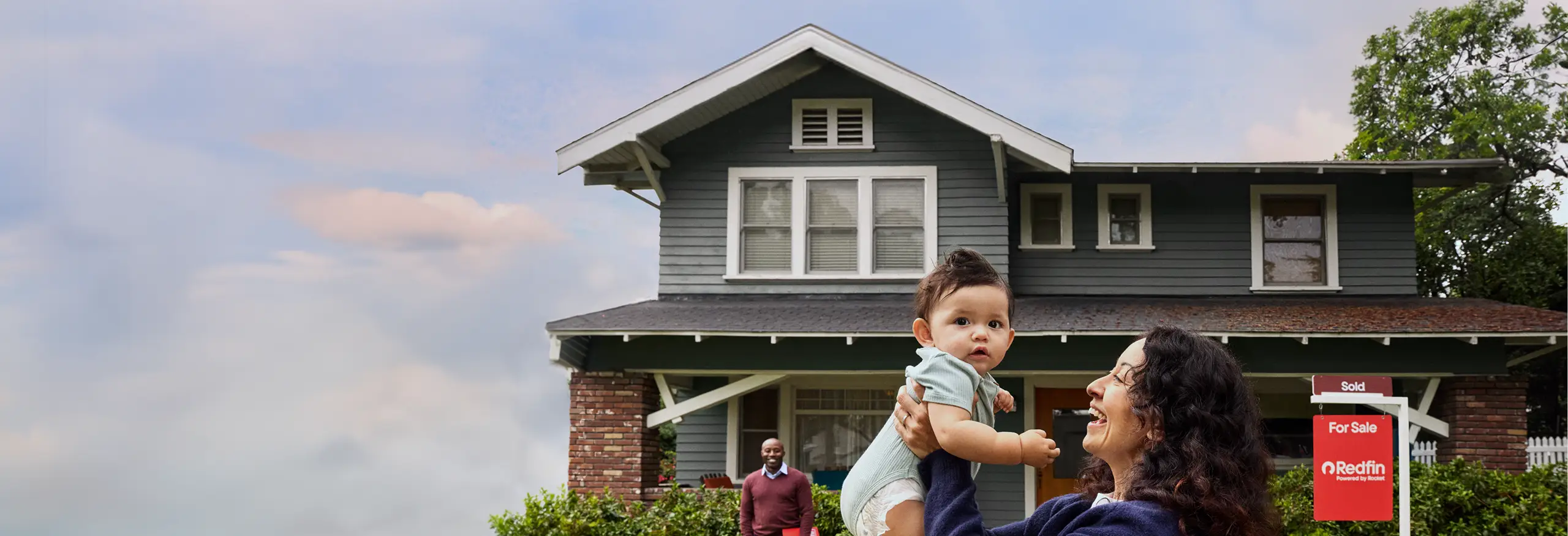 A woman lifting a baby in front of a green two-story house with a Redfin “Sold” sign in the yard while the realtor stands behind them.