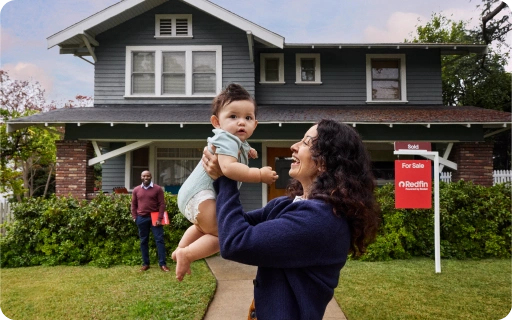A woman lifting a baby in front of a green two-story house with a Redfin “Sold” sign in the yard while the realtor stands behind them.