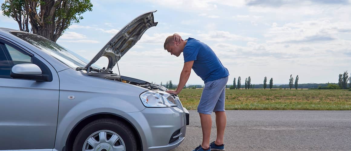 A young man near a broken car with an open hood on the roadside.