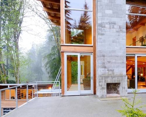 Contemporary home on pine tree wooded lot featuring mirrored picture windows and stone outdoor fireplace.