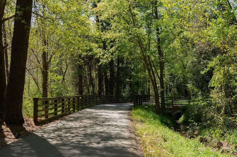 Paved path through a heavily wooded greenway, fencing and drainage ditch flanking the path, North Augusta South Carolina.