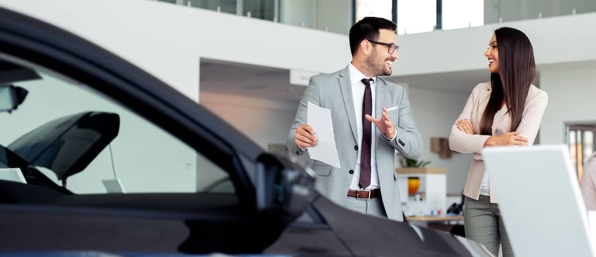 Man in a suit showing a car to a woman and smiling.