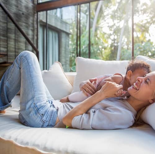 Mother snuggling and smiling with her baby on white couch with large sliding glass doors behind her featuring wooded yard.