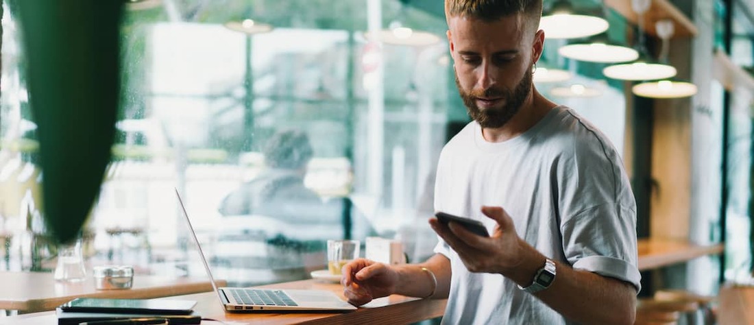 Man looking at cell phone with laptop open at a cafe.