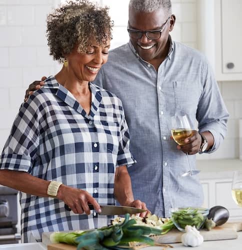Retired couple cooking in kitchen together, smiling and drinking wine.
