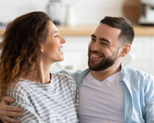 A couple smiling and talking while sitting on the couch in their house.