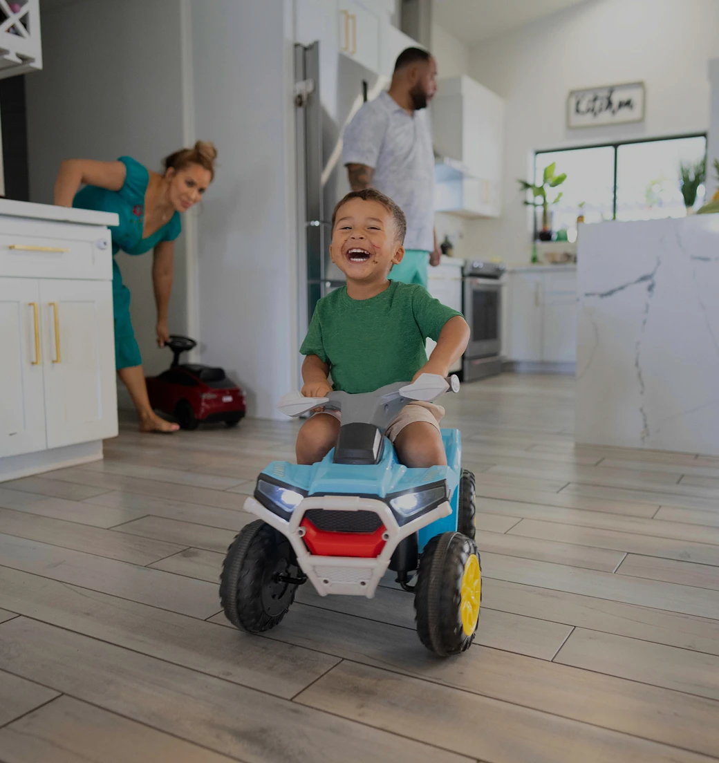 Child laughing while riding his toy car in the kitchen while his parents walk into the room.